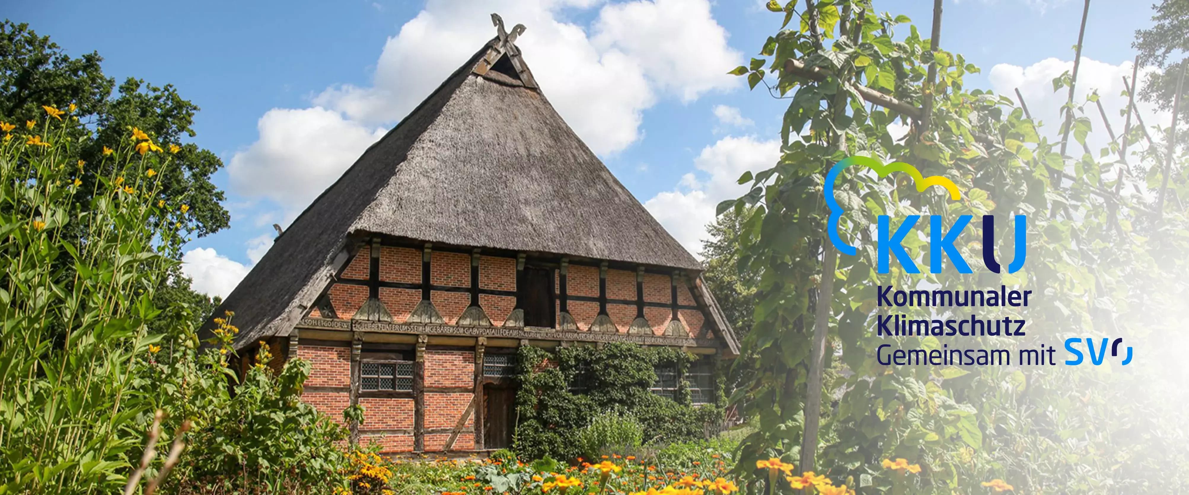 Traditionelles Fachwerkhaus mit Reetdach, umgeben von blühendem Garten und hoher Vegetation, unter blauem Himmel. Rechts im Bild das Logo „KKU – Kommunaler Klimaschutz gemeinsam mit SVO“.