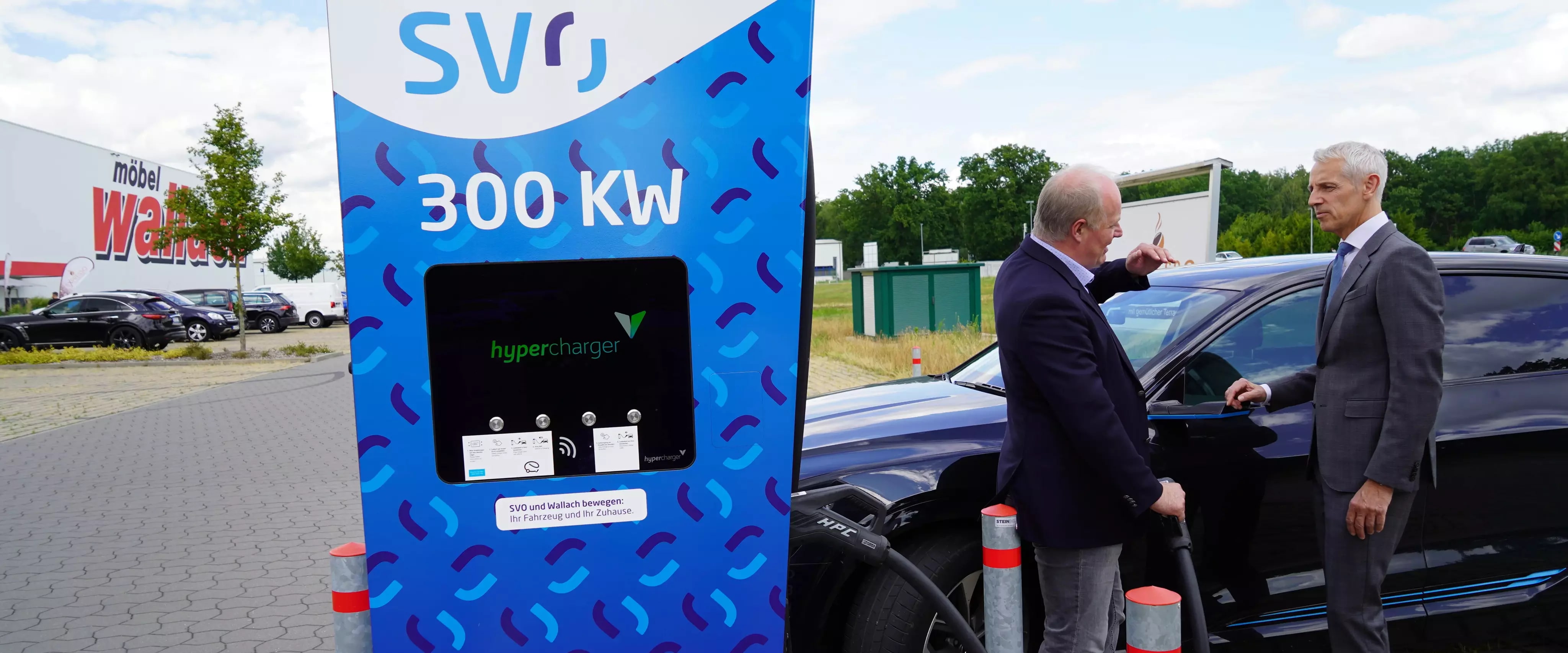 Zwei Männer im Anzug unterhalten sich neben einem Elektroauto an einer 300-kW-Hypercharger-Ladesäule der SVO, im Hintergrund ist ein Möbelhaus mit Parkplatz zu sehen.