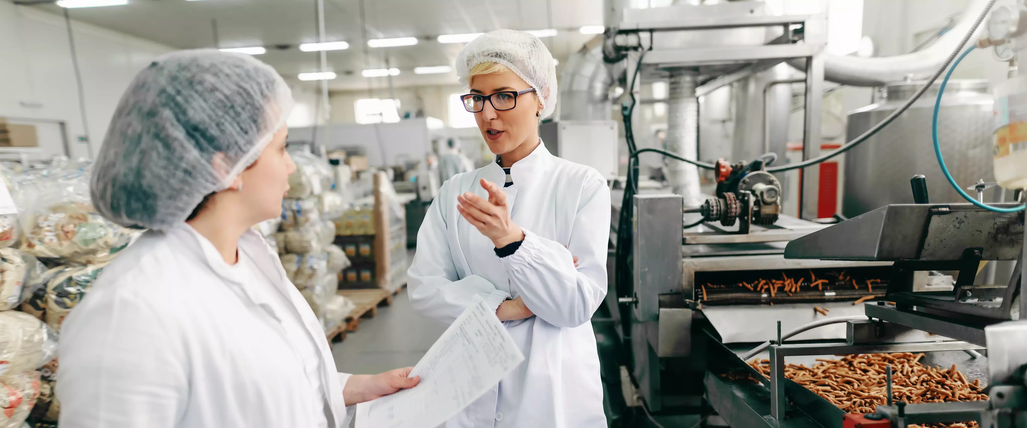 Zwei Frauen in Hygienekleidung und Haarnetzen unterhalten sich in einer Lebensmittelproduktionsanlage; eine hält ein Klemmbrett, im Hintergrund laufen Maschinen und verpackte Waren sind gestapelt.