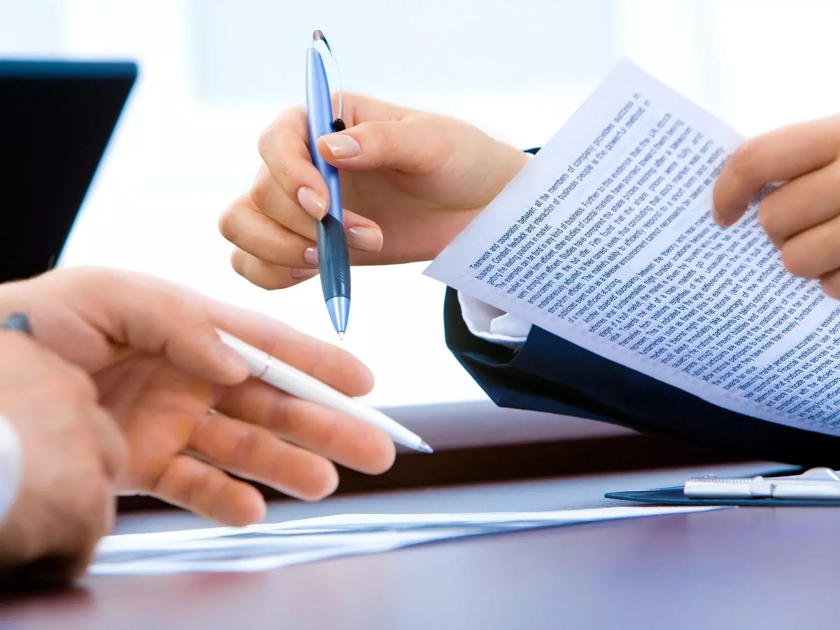 Zwei Personen im Gespräch über ein Dokument. Eine Hand hält ein Blatt Papier mit dichtem Text, während die andere Person mit einem Stift darauf zeigt. Auf dem Tisch liegen weitere Unterlagen und ein Laptop ist im Hintergrund sichtbar.