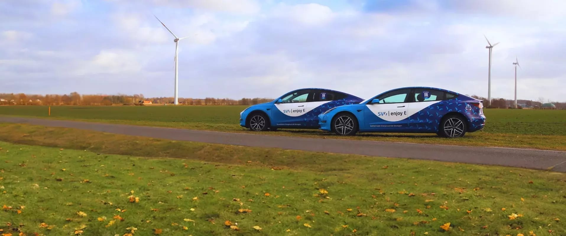 Zwei blau folierte SVO-Elektroautos stehen auf einem Feldweg vor Windrädern.