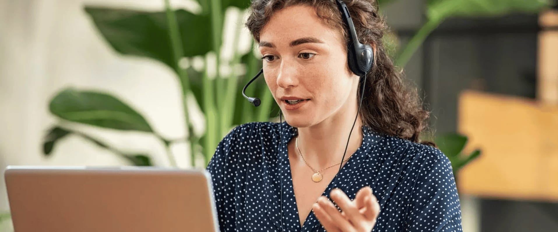 Eine Frau mit Headset sitzt vor einem Laptop und führt ein Online-Gespräch oder einen Kundenservice-Call. Im Hintergrund sind grüne Pflanzen zu sehen.