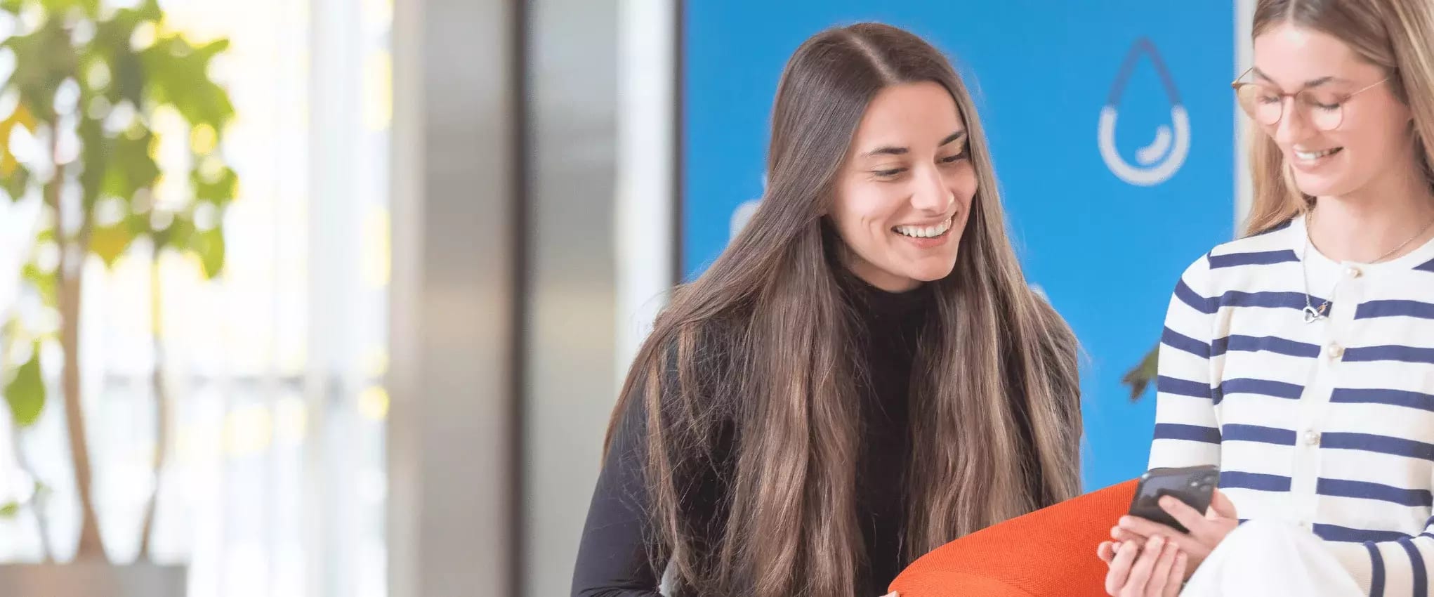 Zwei junge Frauen lächeln und schauen gemeinsam auf ein Smartphone.