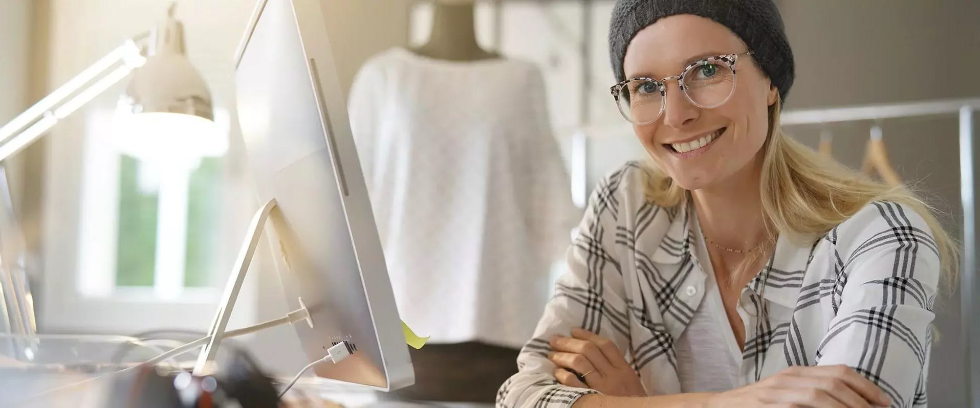 Junge Frau mit Brille und Mütze sitzt lächelnd an einem Schreibtisch vor einem Computerbildschirm in einem hellen, modernen Raum mit Schneiderpuppe im Hintergrund.