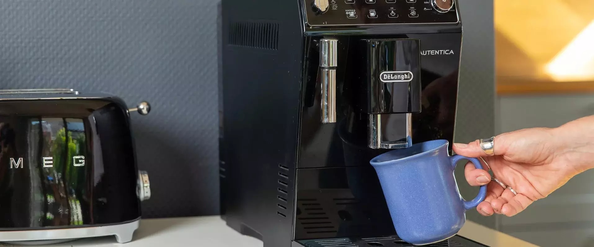 Eine Person hält eine blaue Tasse unter den Auslauf einer schwarzen De’Longhi-Kaffeemaschine. Daneben steht ein schwarzer SMEG-Toaster auf der Küchenzeile.