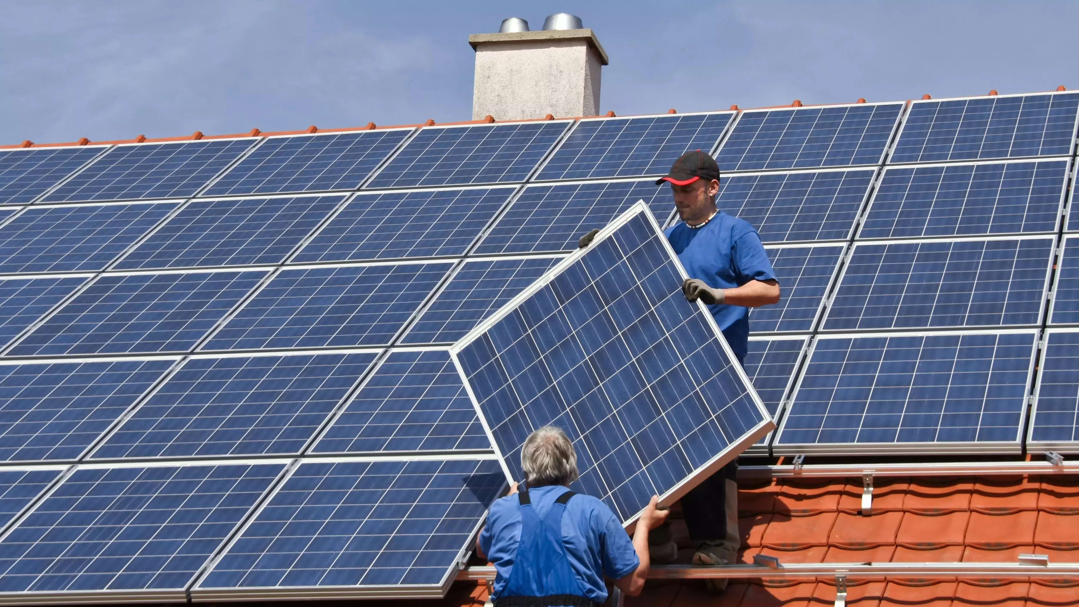 Zwei Techniker installieren Solarpanels auf dem Dach eines Gebäudes bei sonnigem Wetter.