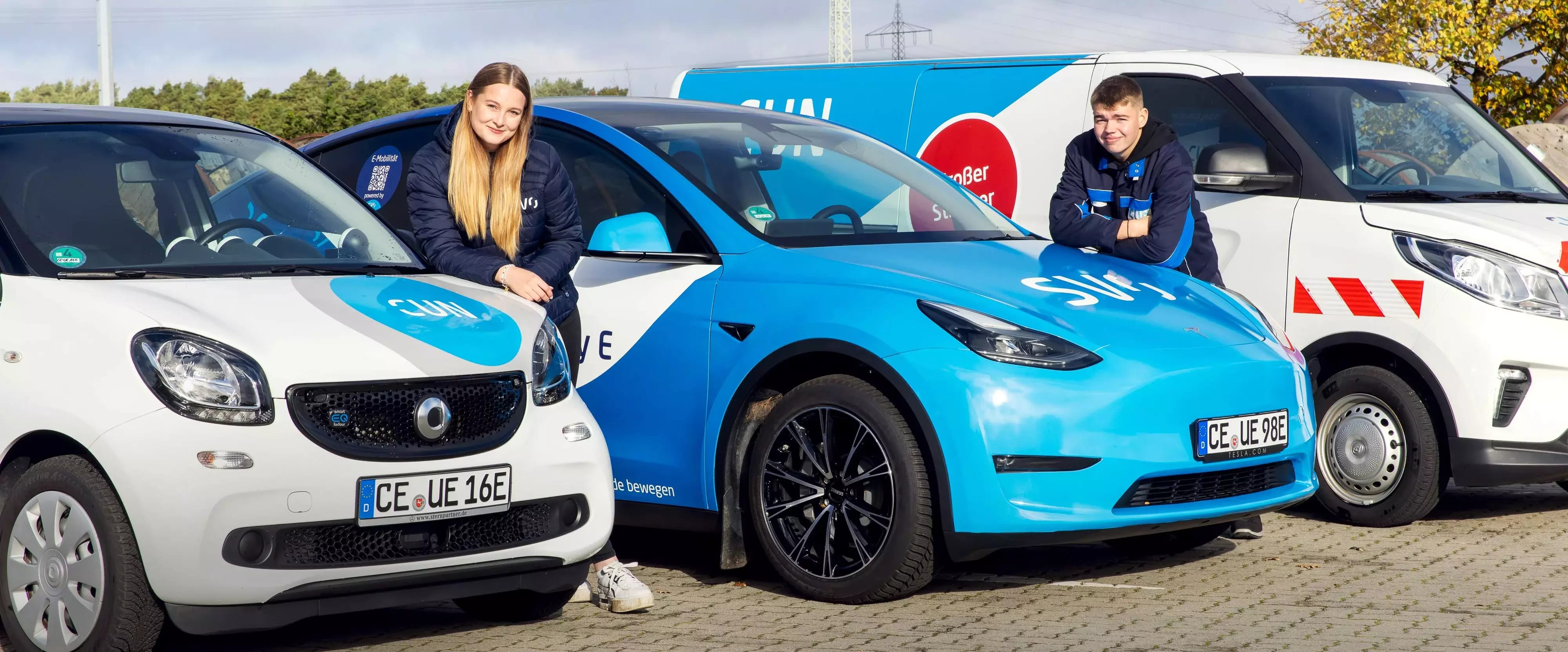 Zwei junge Mitarbeitende der SVO-Gruppe lehnen lächelnd an E-Fahrzeugen mit Firmenlogo – im Hintergrund ein weiteres Servicefahrzeug. Das Bild vermittelt Teamgeist und nachhaltige Mobilität.