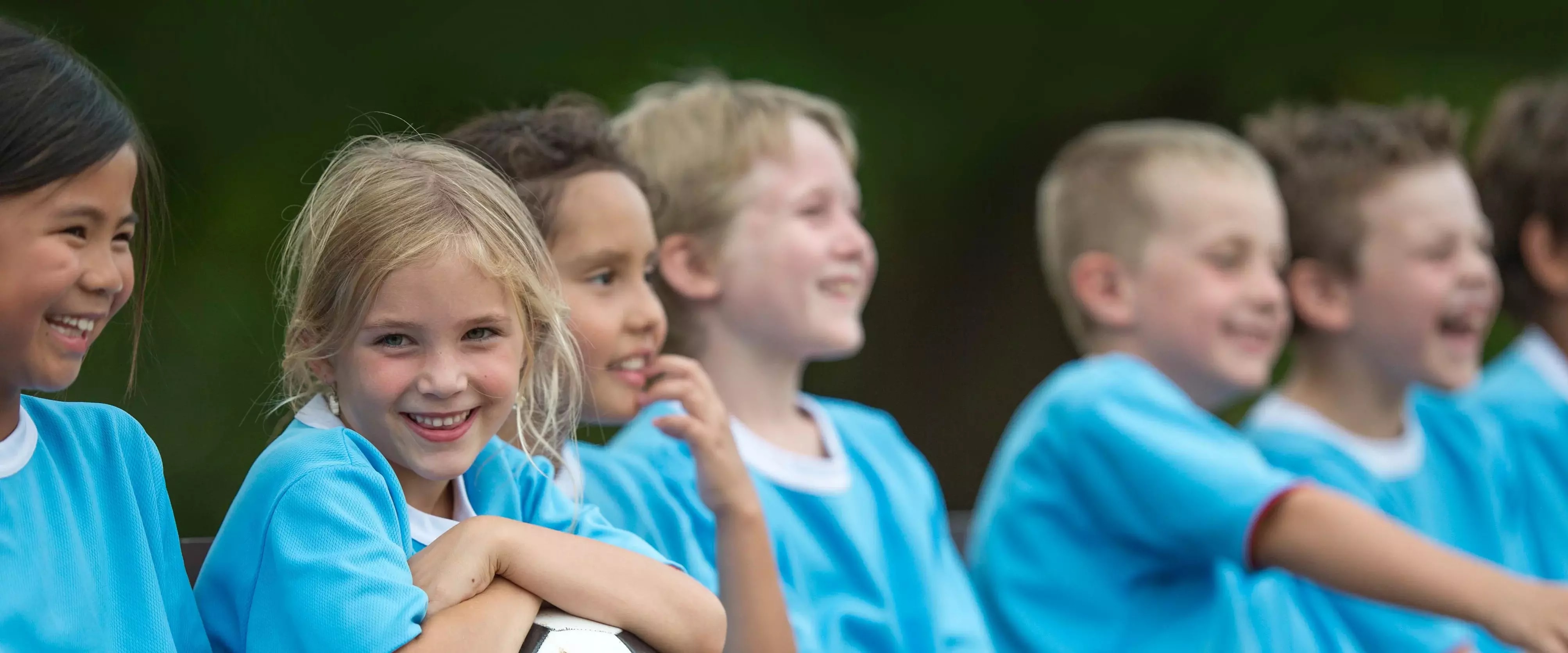 Kinder in blauen Trikots sitzen lachend nebeneinander, ein Mädchen hält einen Fußball im Arm.