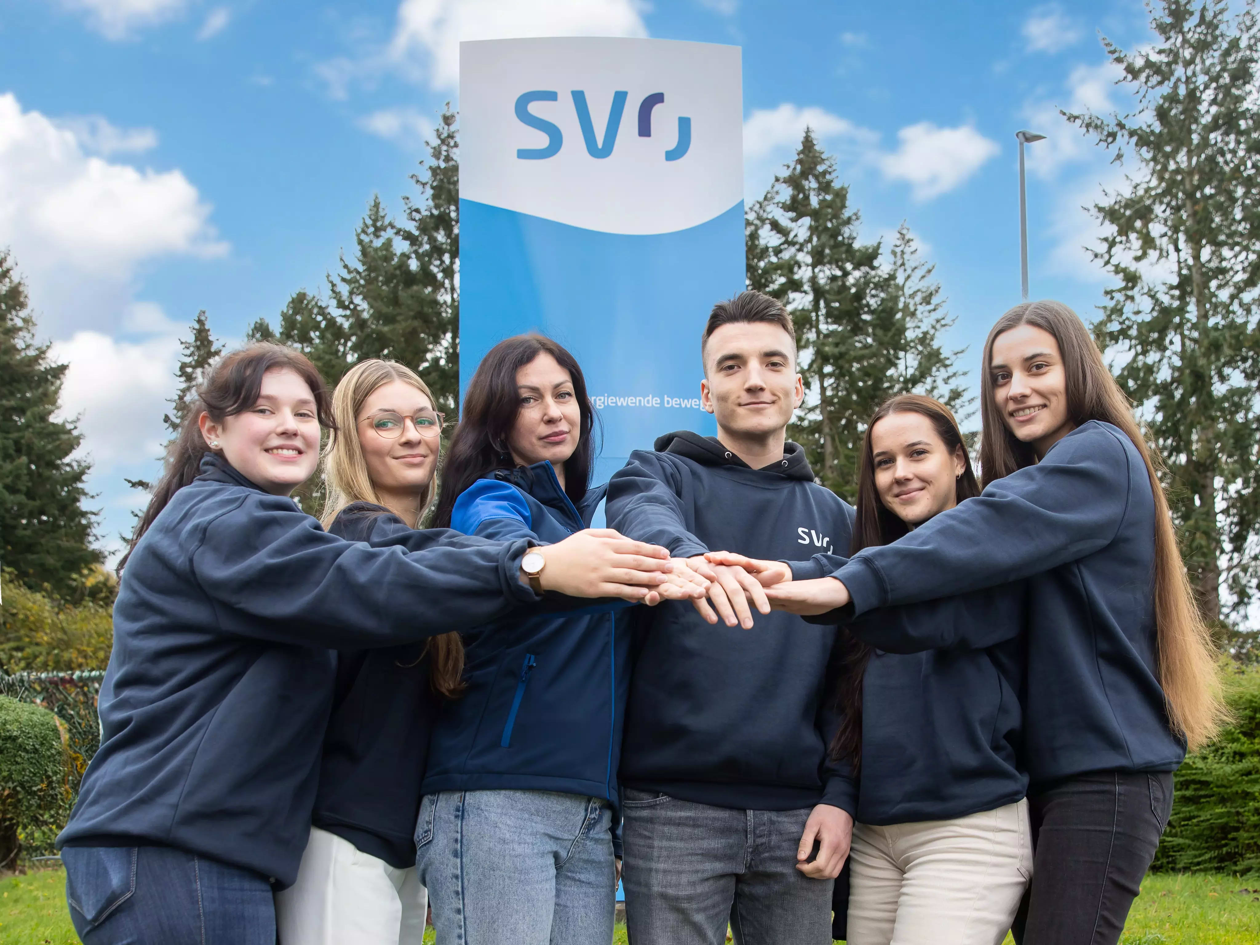 Sechs junge Menschen in dunkelblauen Oberteilen stehen vor einem SVO-Firmenschild und legen motiviert ihre Hände übereinander. Sie lächeln in die Kamera, hinter ihnen sind grüne Bäume und ein blauer Himmel mit einigen Wolken zu sehen.