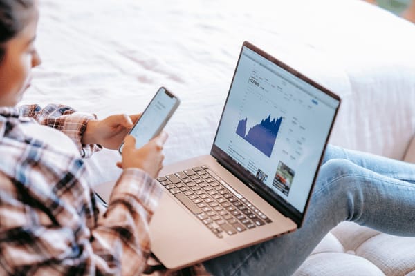 Frau prüft Börsenkurse auf dem Laptop und hält ein Smartphone in der Hand.