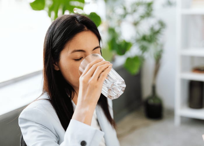 Frau trinkt Wasser aus einem Glas in heller Innenraumumgebung.
