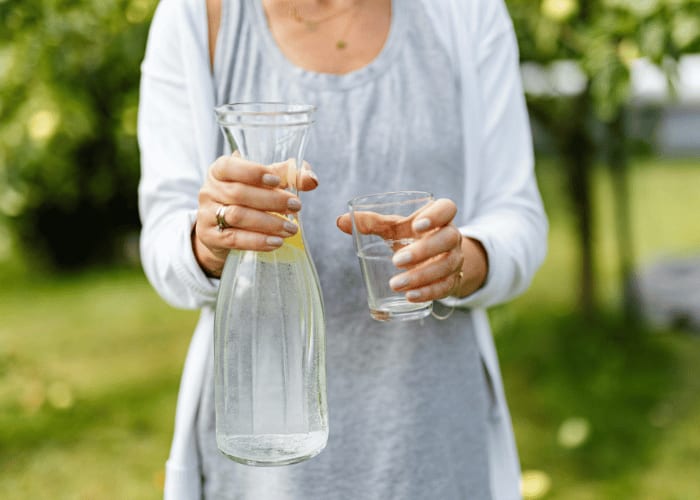 Person hält eine Wasserkaraffe und ein Glas mit frischem Wasser im natürlichen Umfeld.