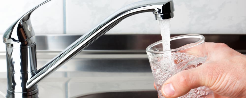Eine Hand hält ein Glas unter einen laufenden Wasserhahn in einer Küchenspüle. Das Glas wird mit klarem Leitungswasser gefüllt.