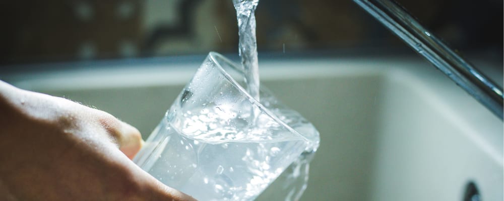 Ein Glas wird schräg unter einen laufenden Wasserhahn gehalten und mit fließendem Leitungswasser gefüllt.