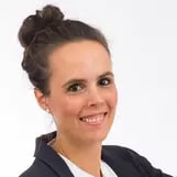 Porträt einer lächelnden Frau mit dunklem Dutt, im Blazer vor hellem Hintergrund.