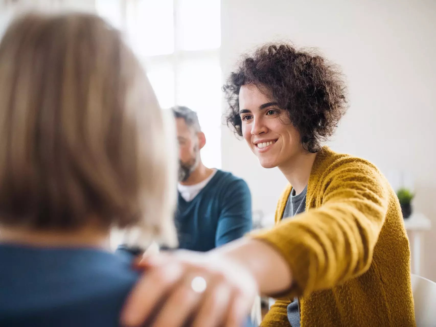 Junge Frau mit lockigem Haar legt tröstend die Hand auf die Schulter einer anderen Person und lächelt.