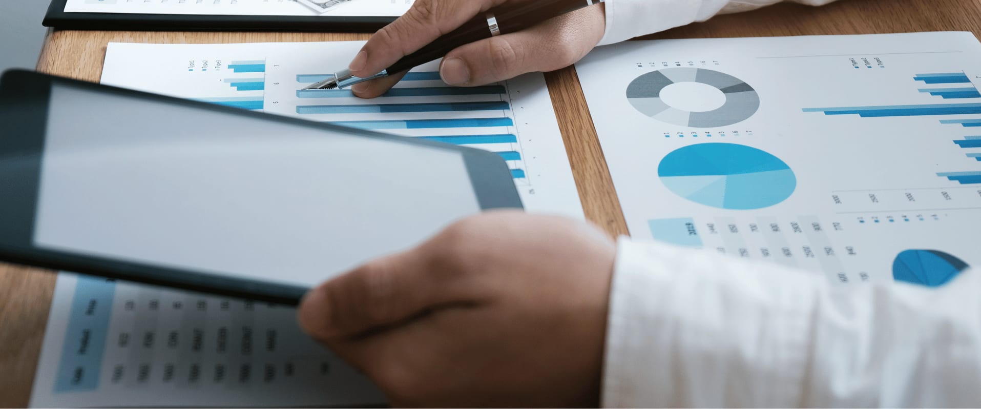 Zwei Hände analysieren Finanzdiagramme mit einem Tablet und einem Stift. Auf dem Tisch liegen verschiedene Ausdrucke mit Balken-, Kreis- und Säulendiagrammen in Blautönen.