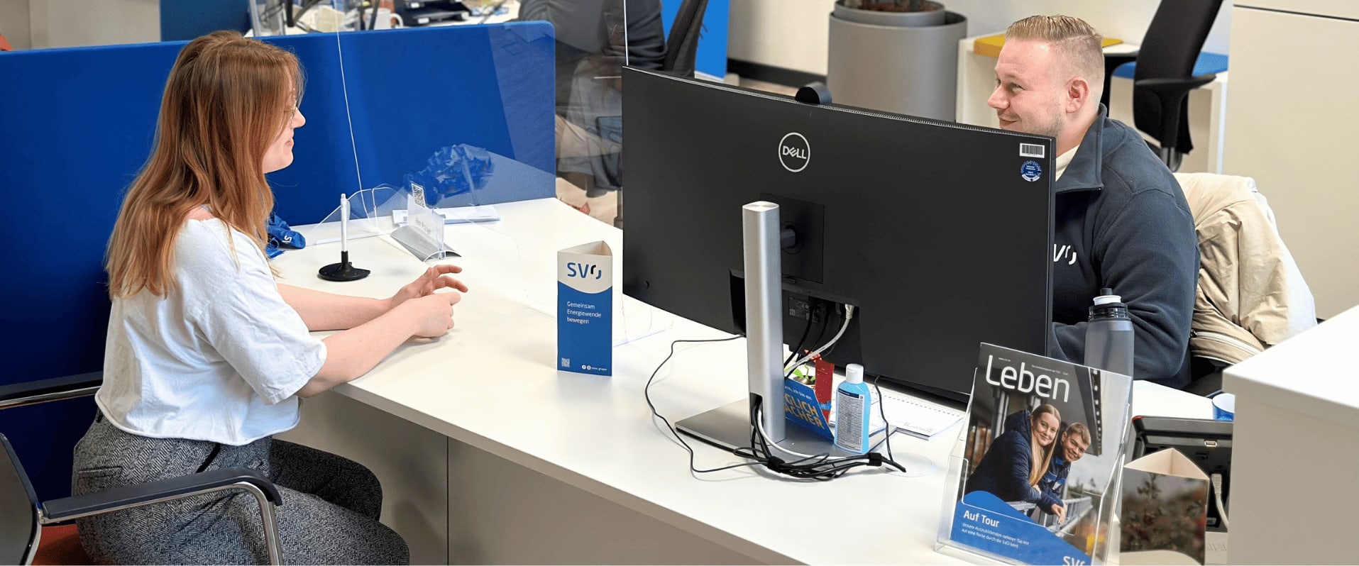 Eine junge Frau sitzt an einem Beratungsschalter der SVO und spricht mit einem Mitarbeiter, der hinter einem Computerbildschirm sitzt. Auf dem Tisch stehen Informationsmaterialien und eine Trennscheibe aus Plexiglas.