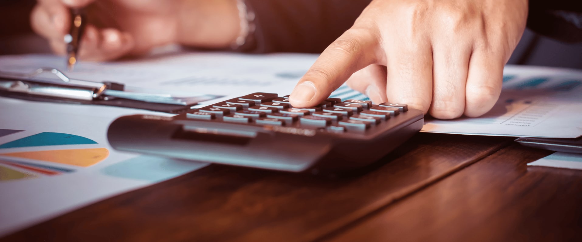 Eine Person tippt auf einem Taschenrechner, umgeben von Dokumenten mit Diagrammen und einem Stift auf einem Schreibtisch.