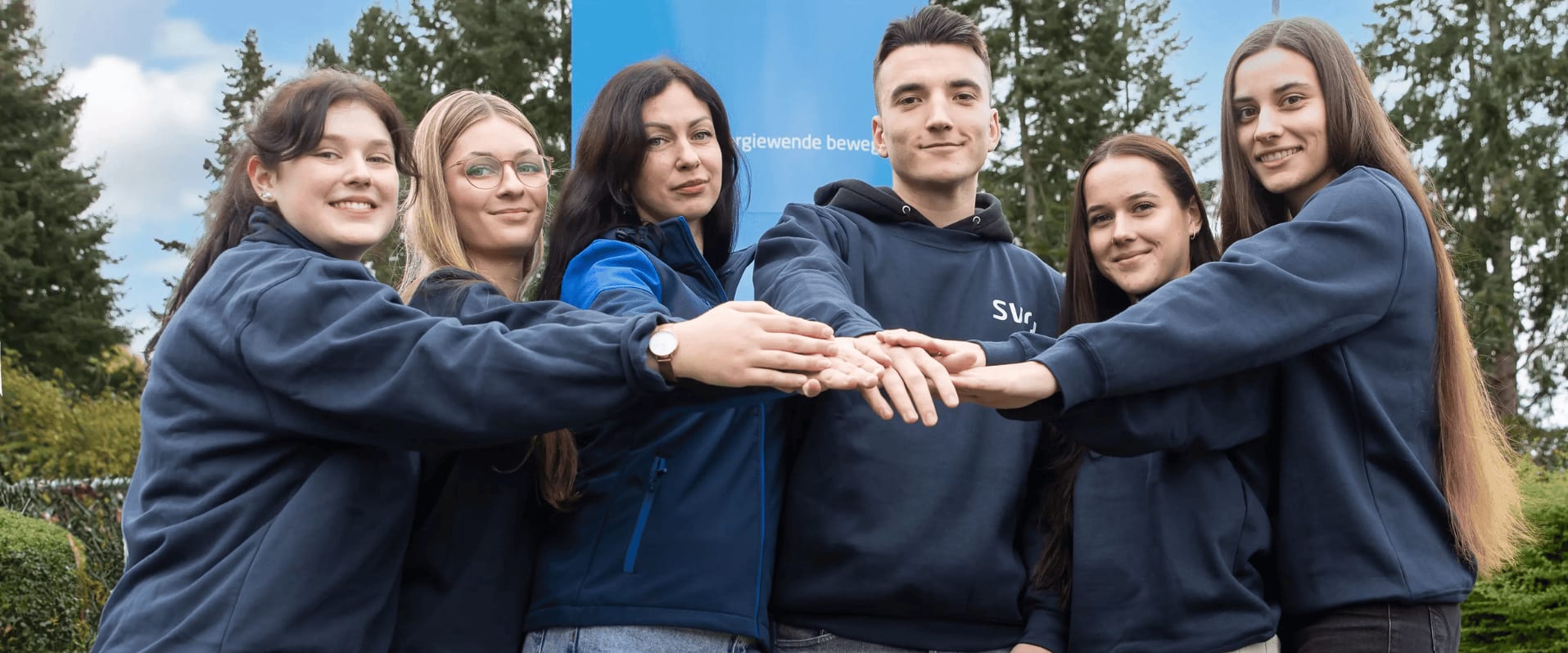 Sechs junge Menschen in dunkelblauen Oberteilen stehen vor einem SVR-Firmenschild und legen motiviert ihre Hände übereinander. Sie lächeln in die Kamera, hinter ihnen sind grüne Bäume und ein blauer Himmel mit einigen Wolken zu sehen.