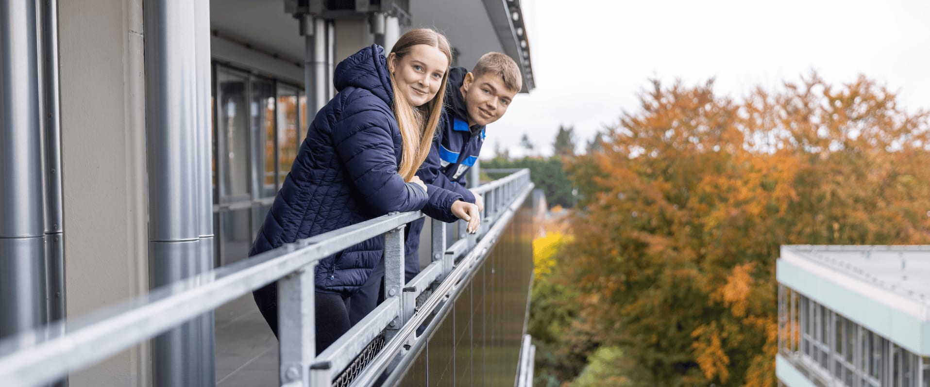 Zwei junge Menschen lehnen sich über ein Balkongeländer eines modernen Gebäudes. Beide tragen blaue Jacken und lächeln in die Kamera. Im Hintergrund sind herbstlich gefärbte Bäume mit orangefarbenem Laub zu sehen.