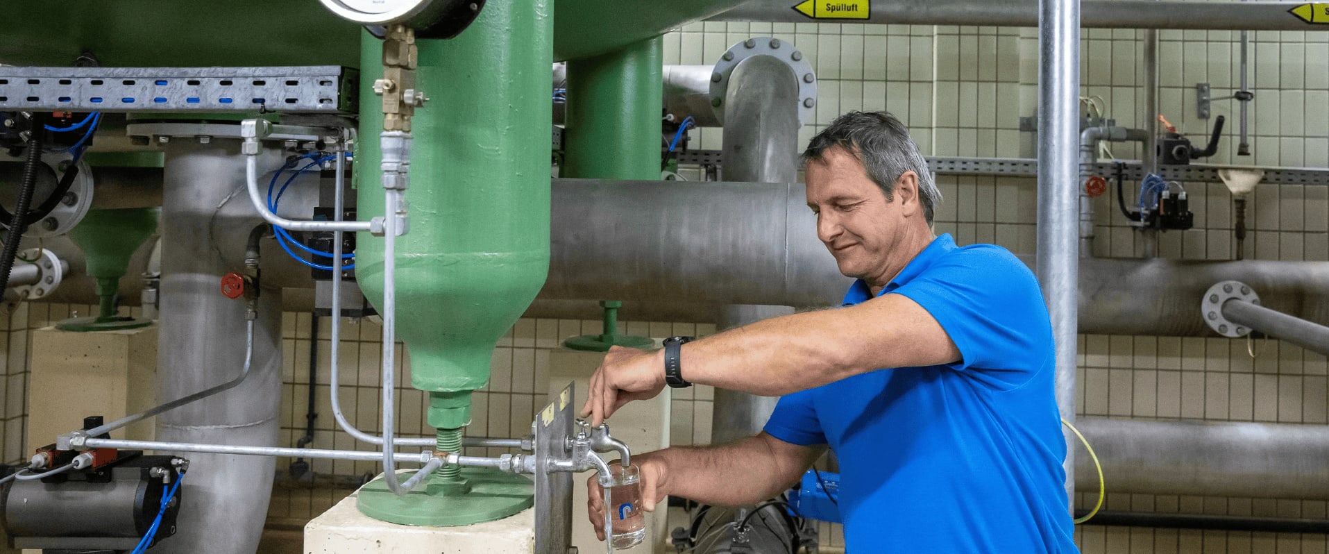 Ein Mann in blauer Arbeitskleidung zapft in einer technischen Anlage Wasser aus einem Hahn in ein Glas; im Hintergrund sind große grüne und silberne Rohrsysteme zu sehen.