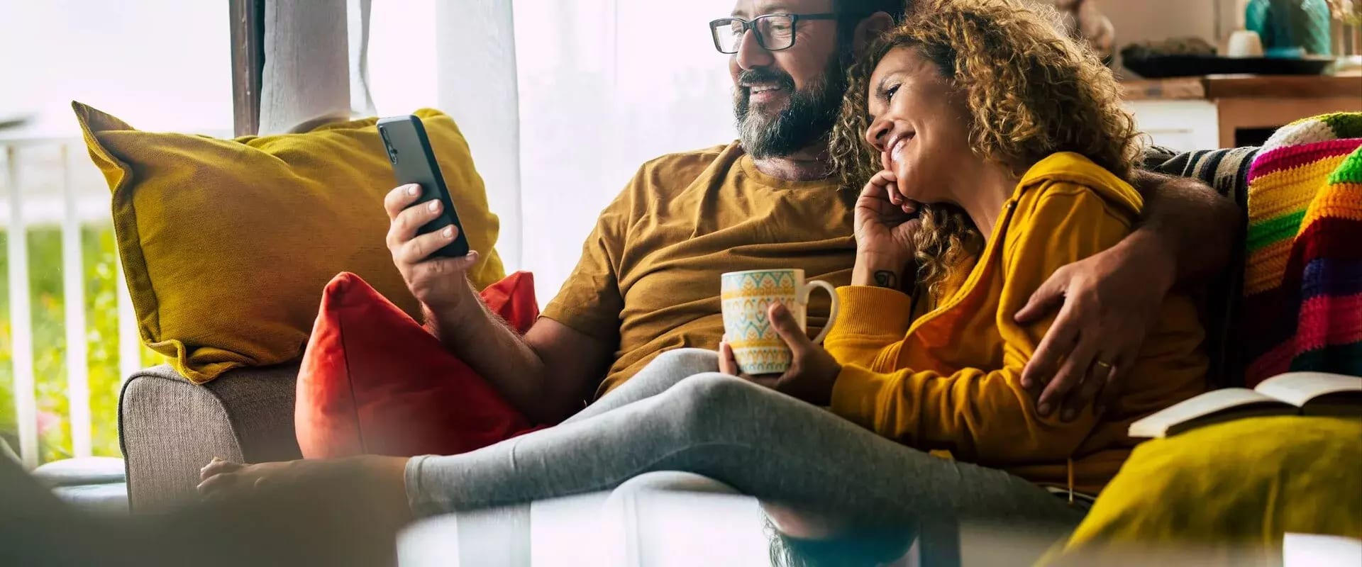 Ein Paar sitzt entspannt auf dem Sofa, schaut gemeinsam auf ein Smartphone und genießt eine heiße Tasse Kaffee.