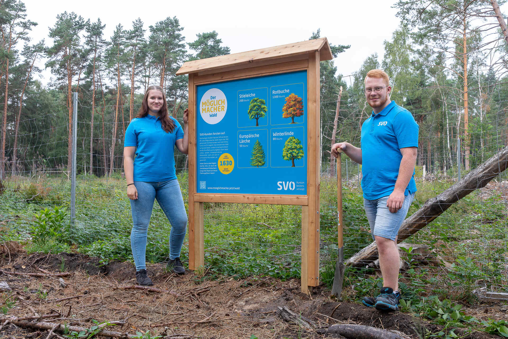 Zwei junge Menschen in blauen SVO-Shirts stehen neben einem Schild im Wald, das über gepflanzte Bäume im „Möglichmacher-Wald“ informiert.
