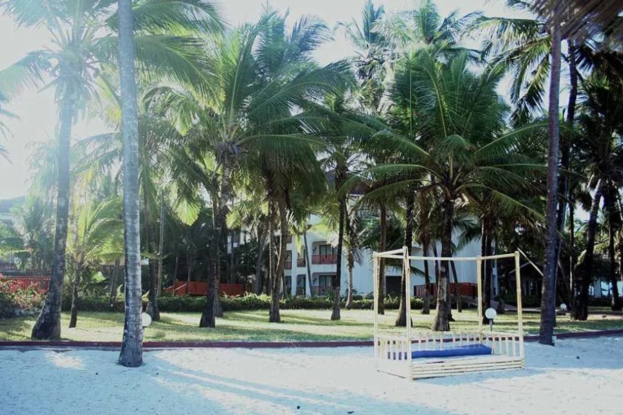 Palm-lined beach at a Diani resort, with a white hotel building and a wooden daybed on the sand
