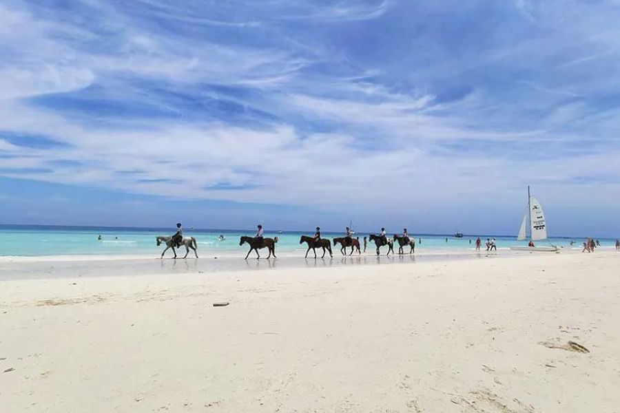 Tourists horseback riding along a white sand beach in Zanzibar, with a sailboat nearby
