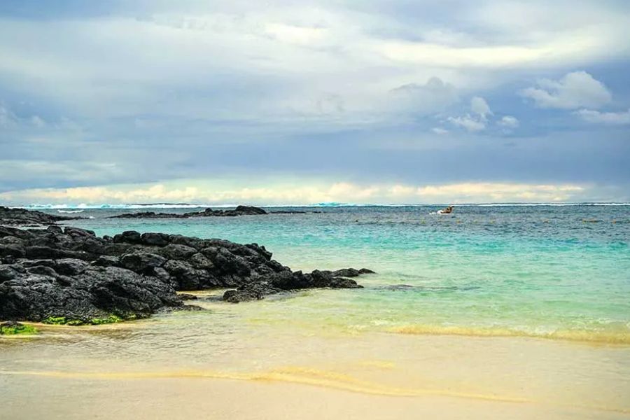 Black volcanic rocks along a quiet Mauritius beach, calm turquoise water under cloudy sky