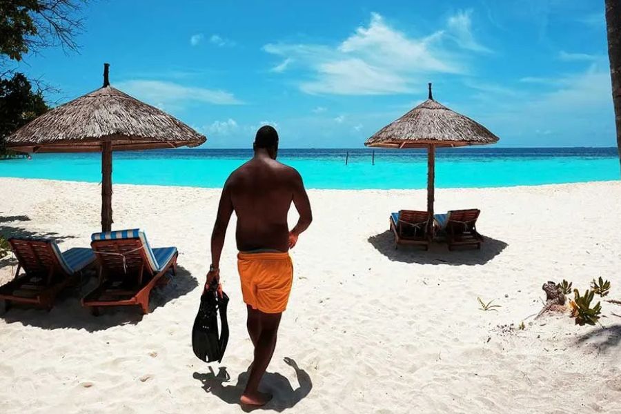 Man walking toward the water with snorkel fins, thatched umbrellas on a Maldives beach