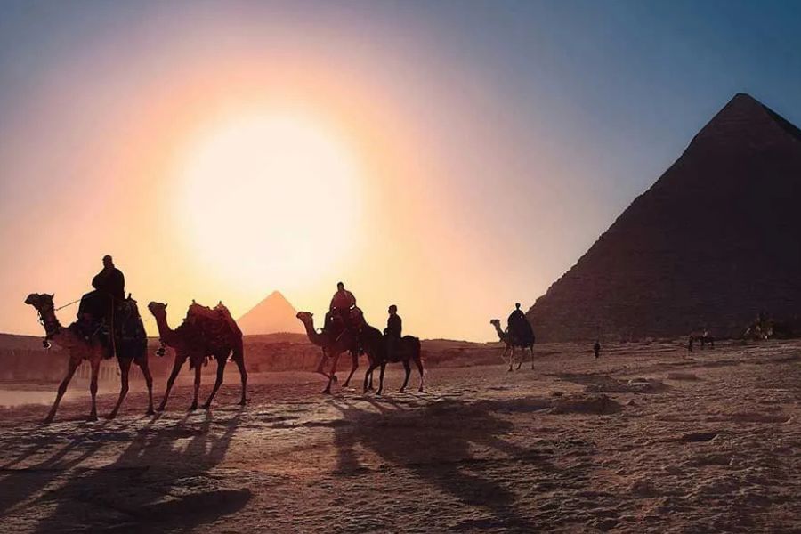 Silhouettes of camels and riders walking toward pyramids at sunset