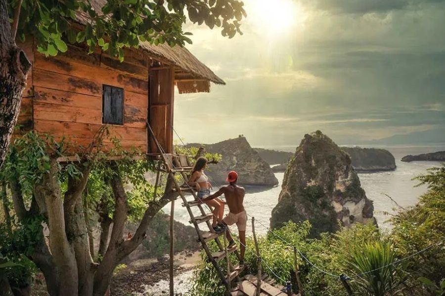 Couple climbing a ladder to a wooden treehouse overlooking rocky cliffs and the sea