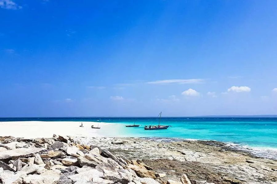 Dhow boats anchored near a rocky shoreline with white sand and turquoise water