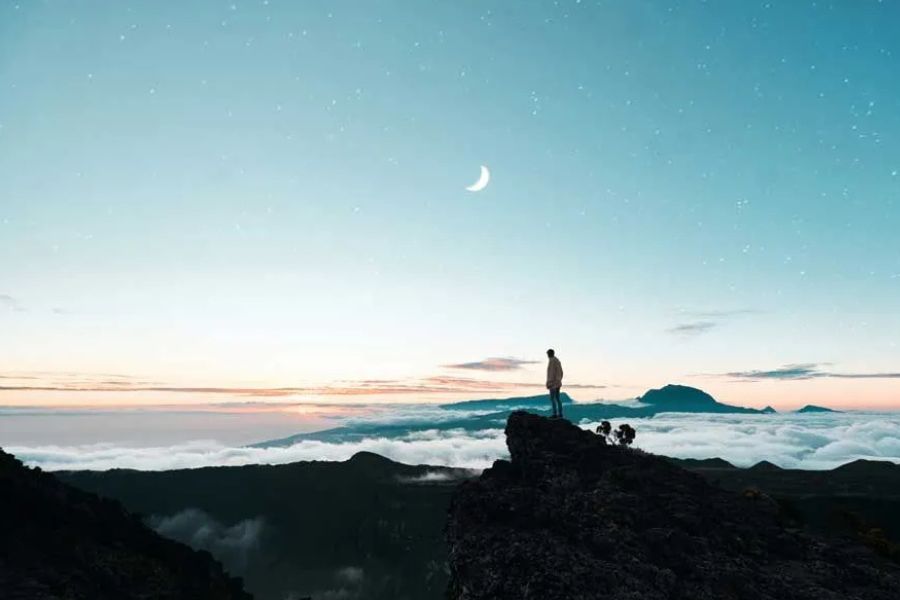 Person standing on a rocky peak above clouds under a starry night sky with crescent moon
