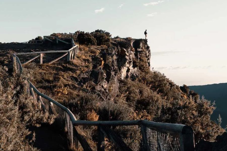 Person standing on a rocky cliff edge along a wooden fence hiking trail
