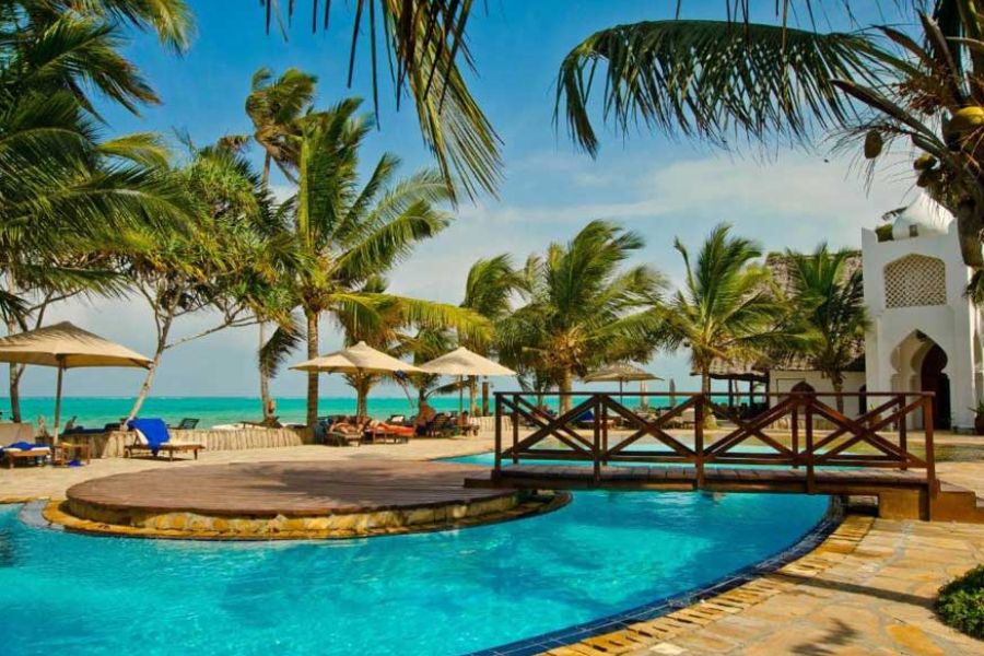 Resort swimming pool with a wooden bridge, sun umbrellas, and ocean view