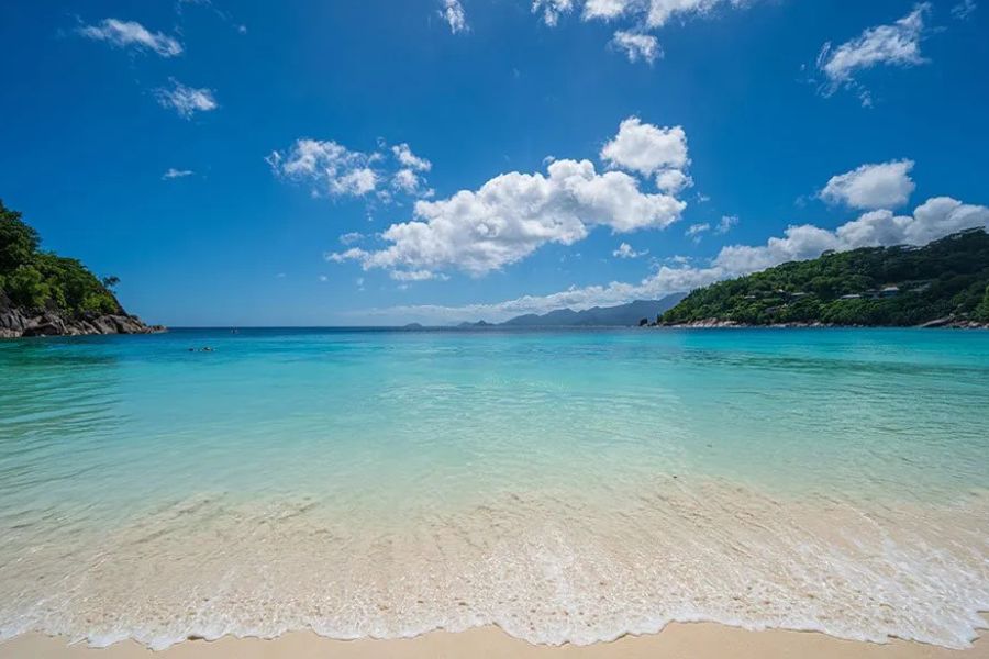 Calm, shallow turquoise bay and white sand beach in the Seychelles, with green hills in the background