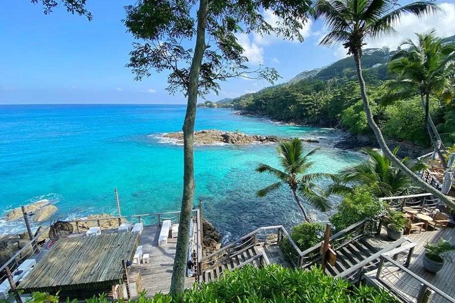 Wooden steps lead down a palm-lined granite cliff to a turquoise cove along the Seychelles coastline