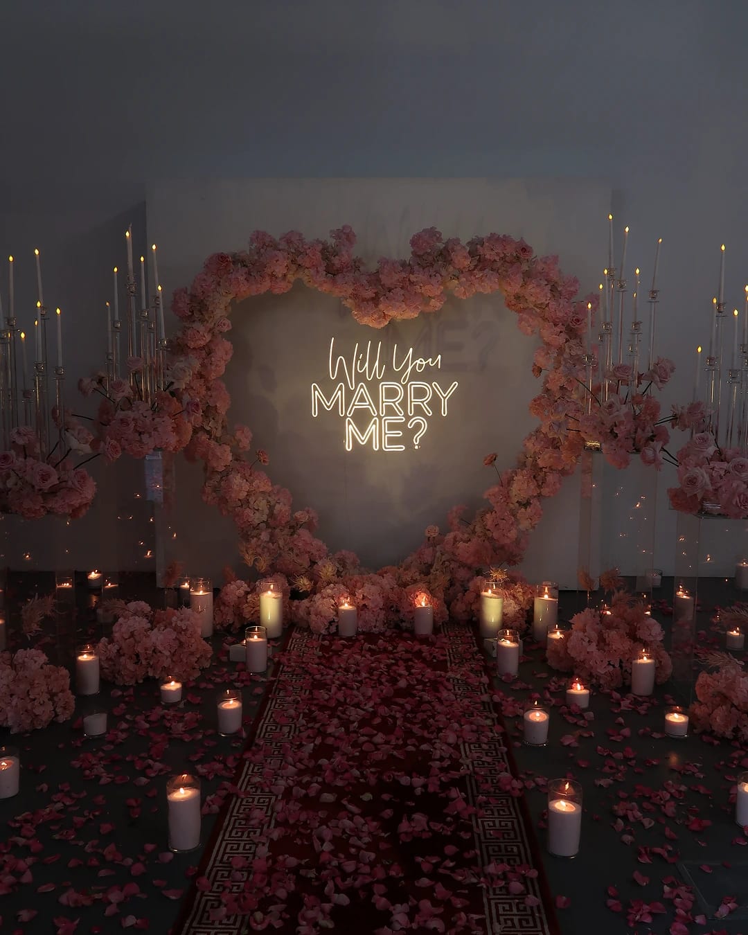Romantic marriage proposal setup with floral heart backdrop, candles and personalised signage