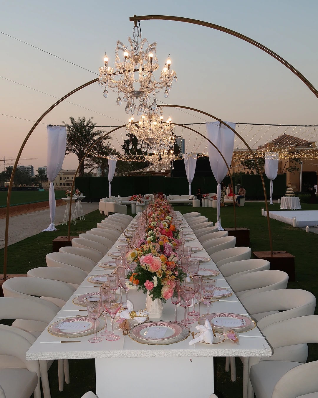 Outdoor luxury long table dinner setup with chandeliers, floral décor and premium seating arrangement