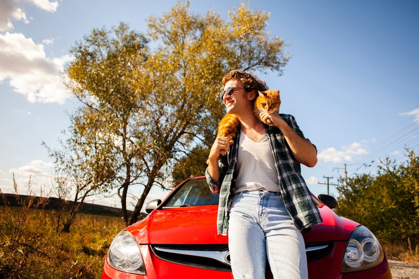 Hombre con un gato en su espalda, de pie apoyado en un auto rojo, con un árbol de fondo.