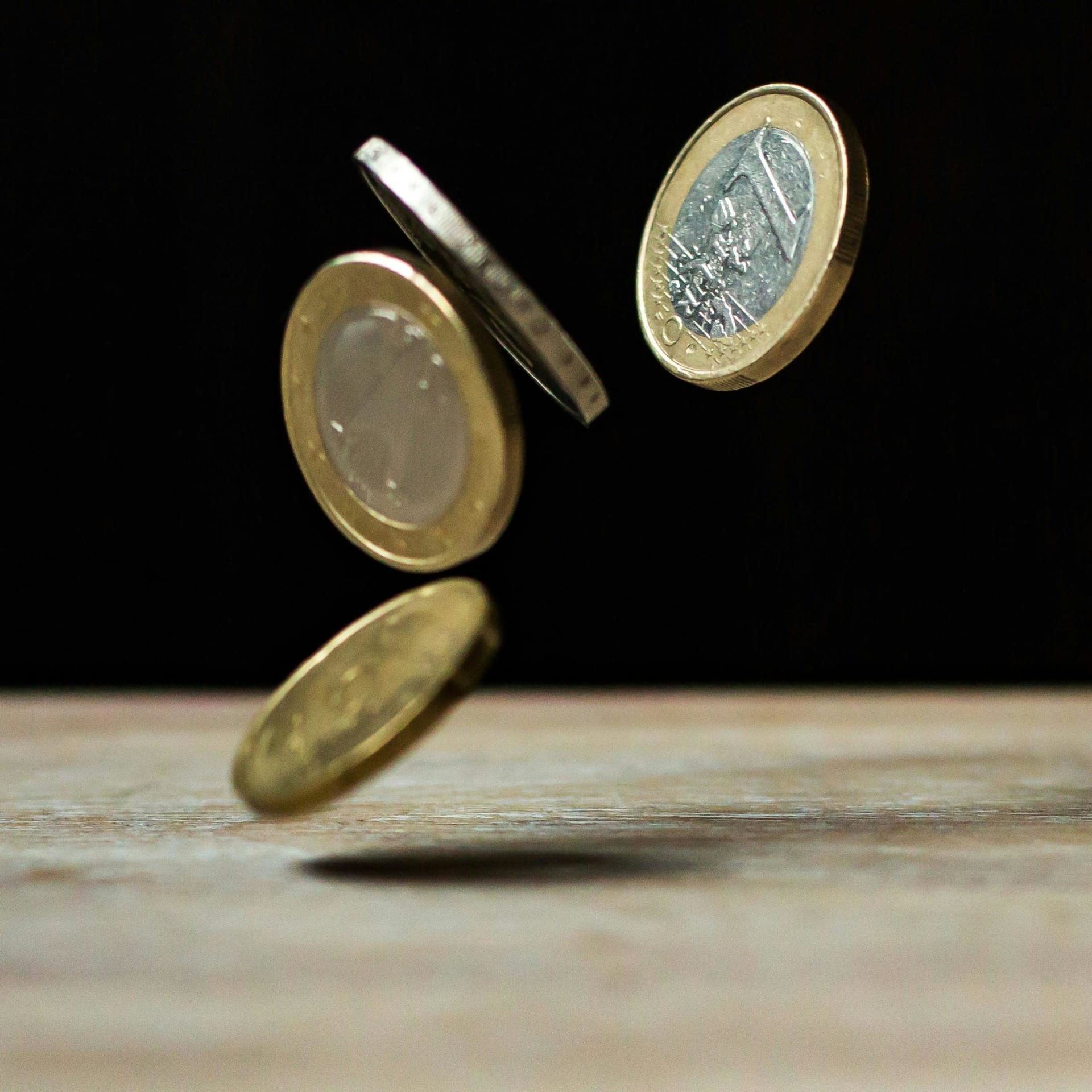 Three Euro coins suspended in mid-air against a dark background, with one leaning on another on a wooden surface. The coins display various angles, revealing different faces, with one coin prominently showing the '1 Euro' denomination. The lighting highlights their metallic texture.