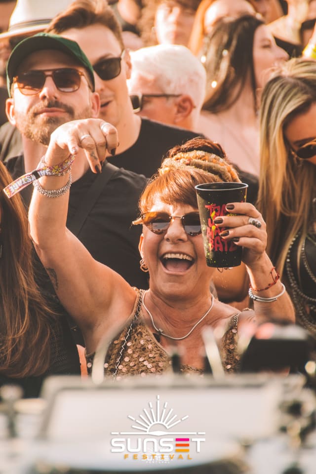 Participante sorri e levanta o braço e o copo do festival em meio aos amigos na pista.