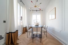 Dining room with a black round table, six chairs, designer chandelier, and tall curtains.