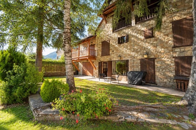 Stone facade of the house with front garden, trees, and a small terrace with chairs.
