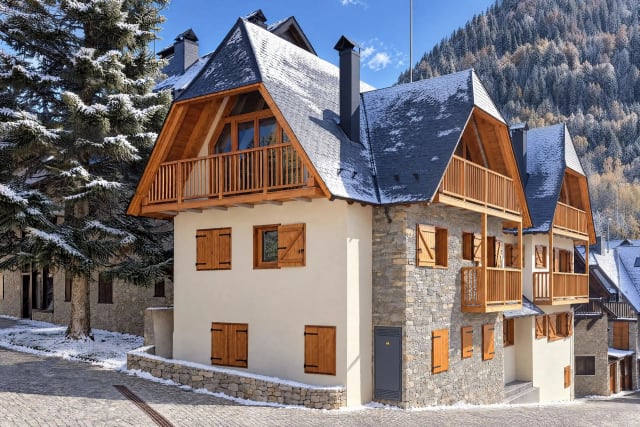 Side view of the building from the cobbled square, with light snow on the slate roofs, a tall fir tree and the snowy mountain behind.