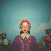 Mulher vestida de roxo, olhos fechados e coroa de flores na cabeça. Em pé num campo florido sob um céu azulado. Imagem gerada com IA.