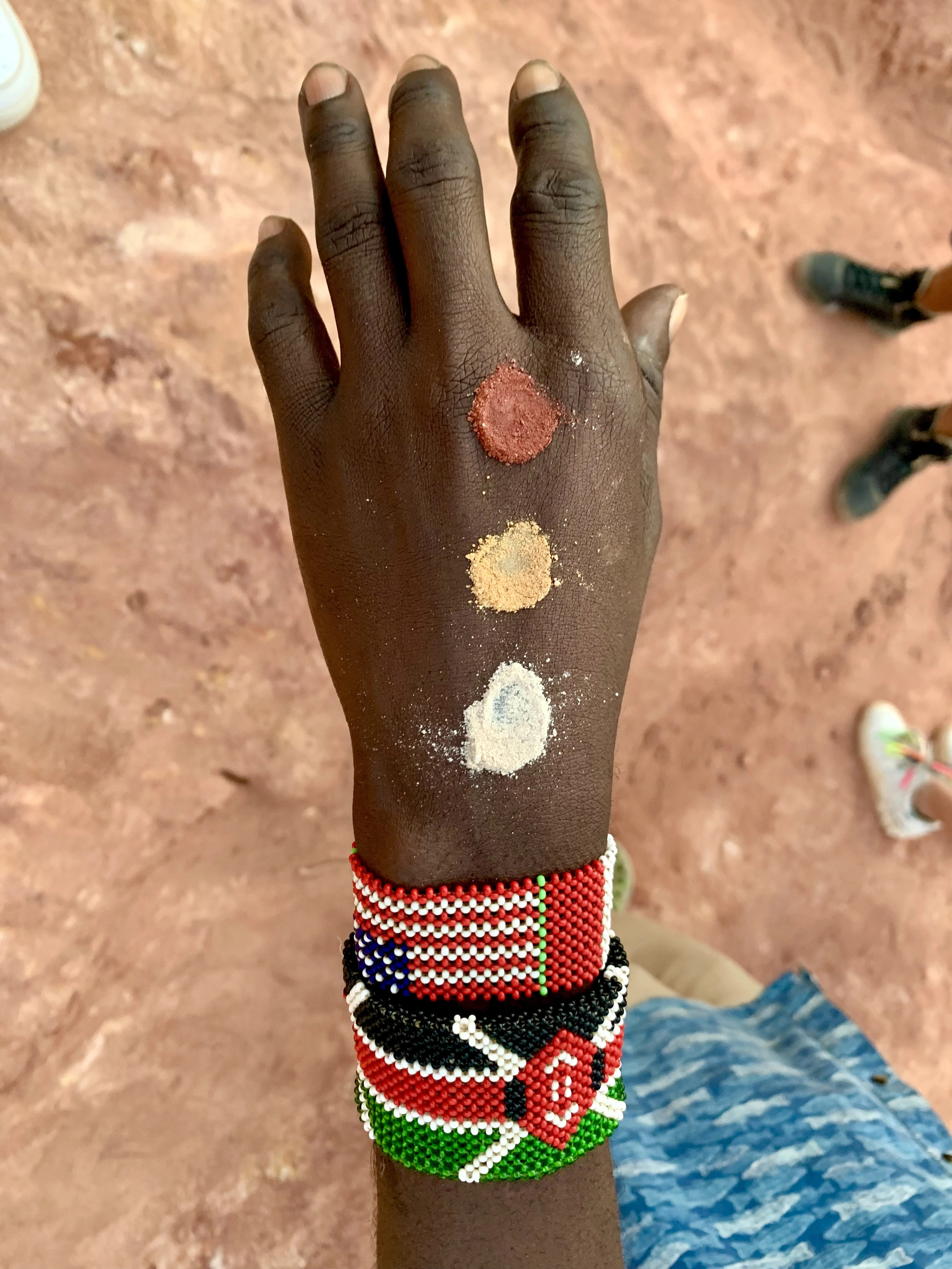 Una di un local mostra tre piccoli cumuli di pigmenti di terra di diversi colori (rosso, ocra, bianco) e due braccialetti colorati con perline ai polsi, sullo sfondo di terra rossa.